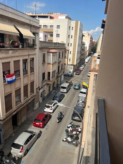The image depicts a street scene in Palma, Spain, viewed from an elevated position, likely a balcony. The street is lined with buildings of varying heights and architectural styles, suggesting an urban environment. The time of day appears to be daytime, indicated by the bright sunlight casting shadows. The weather seems clear and sunny.

Several vehicles are parked along the street, including cars and motorcycles. Among the cars, a red hatchback and a white sedan are prominently visible. The motorcycles are parked in a group, some of them appearing to be sport bikes. Blue parking lines are visible on the road surface, denoting designated parking spots.

The buildings exhibit features such as balconies, awnings, and windows. Some of the buildings have signage. On one of the buildings, the word "HOTEL" is visible in large letters, along with the word "ALUX". Another sign on a shop appears to say "ELENA". A flag, appearing to be the flag of Paraguay, is hanging from a balcony. The overall impression is of a residential and commercial street in a Spanish city. There are no people clearly visible in the image, but the presence of parked vehicles and an active street suggests it is a populated area.
