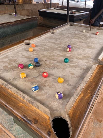 The image shows a person playing billiards on a worn pool table. The table itself appears aged, with a patterned felt surface and a wooden frame. Several billiard balls are scattered across the table, in various states of play. Some are solid colors, while others have stripes and numbers, typical of a game of pool. A white cue ball is visible near the center of the table.
In the background, part of a white vehicle is visible, suggesting an outdoor or semi-outdoor setting. The environment seems to be a bar or a casual recreational area, possibly in Barakin, Nigeria, given the provided location context. The lighting appears to be natural daylight, casting shadows and indicating it's daytime.
The focus is on the game of billiards and the individual playing. The player's arm and hand are visible, holding a cue stick, poised to strike the cue ball. The scene conveys a relaxed atmosphere, common in social settings where games like pool are played. There is no visible text in the image.