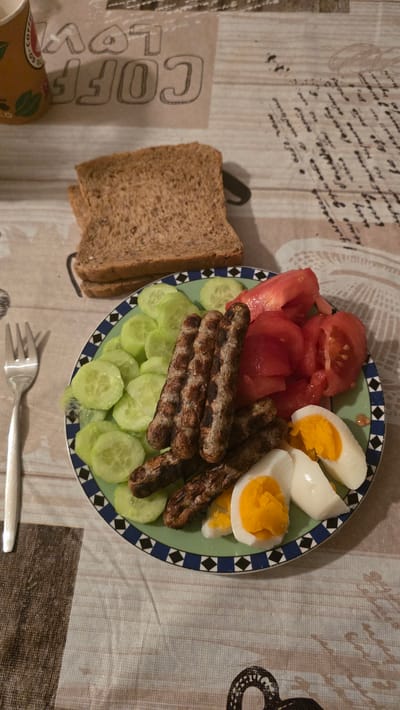 The image shows a plate of food on a patterned tablecloth. The plate contains sliced tomatoes, cucumber, cevapcici, and hard-boiled eggs. Next to the plate, there are slices of brown bread and a fork. There is a coffee cup visible in the lower left corner of the image. There is no indication that this image was taken in Zirndorf, Germany, or any information about people, subjects, or actions.