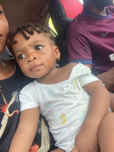 A baby with braided hair and gold hoop earrings lies sideways, looking at the camera. The baby wears a white short-sleeved onesie with dinosaur patterns. An adult wears a maroon Nike shirt and supports the baby. The baby lies on a patterned black and white fabric with red accents. The setting appears to be inside a car, judging by the red car seat and interior details visible in the background.