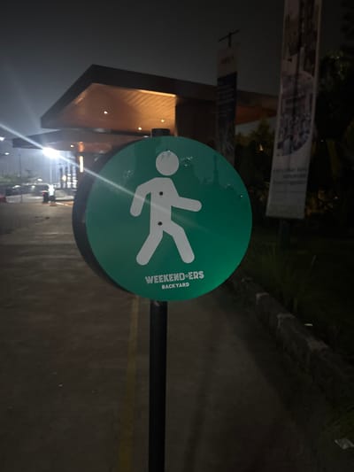 A circular green sign with a white pedestrian icon and the words "WEEKENDERS BACKYARD" is prominently displayed at night in Babakan Madang, Indonesia. The sign is mounted on a dark pole. In the background, a modern building with a lit roof canopy is visible, along with banners and vegetation. The ground appears to be paved. The overall scene suggests an outdoor area likely associated with an event or venue.