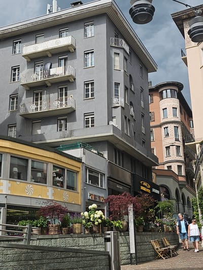 The image depicts a street scene in St. Moritz, Switzerland, featuring a blend of architectural styles. On the left, a grey, multi-story building with numerous windows and white-railed balconies dominates the foreground. Below this building, a yellow facade with large windows displays signs for "Pizza La Stalla Ristorante." In front of the yellow building, a row of potted plants, including a Japanese maple, hydrangeas, and lavender, are arranged on a stone wall.
To the right of the grey building, a distinctive round, pinkish-orange building with ornate white trim and arched windows stands prominently. Further to the right, another building with arches and dark grey facades is visible, housing shops with signage. Two individuals, a man and a woman, are walking on the cobblestone sidewalk, with the man wearing a blue vest and shorts, and the woman in a white top and shorts. Two wooden chairs are positioned on the sidewalk in front of the buildings. The sky is overcast with dramatic clouds. Overhead, two large, industrial-looking light fixtures are suspended.