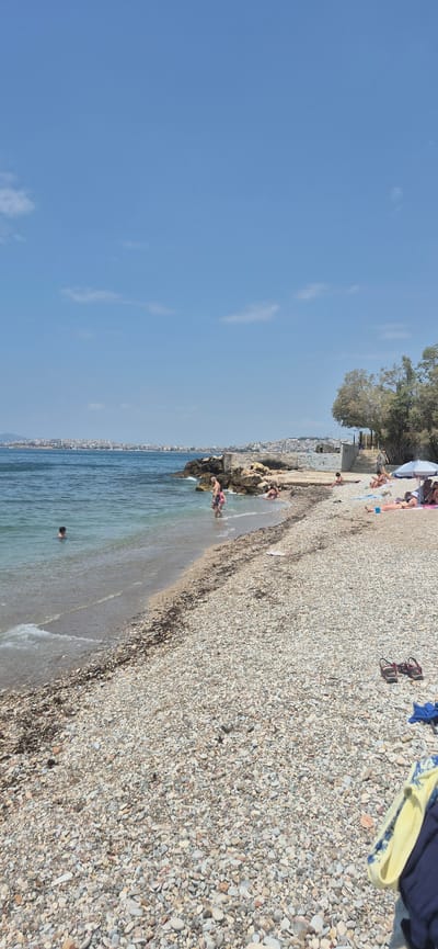 The image depicts a scenic view of a pebble beach in Paleo Faliro, Greece, with the city skyline visible in the distance across the shimmering blue sea. The sky is a clear, bright blue with a few wispy clouds. On the beach, several people are enjoying the sunny day. In the foreground, the pebble-strewn shore is evident, with a few pieces of dark seaweed scattered along the waterline.
Further down the beach, a group of individuals are relaxing. Some are lying on towels, sunbathing, while others are seated, enjoying the atmosphere. A white umbrella provides shade for some of the beachgoers. Closer to the water, a man and a child are wading in the shallows. To the left, a young boy is swimming in the sea. On the right side of the frame, a pair of sandals and some folded clothing are placed on the pebbles. The natural rocky formations and vegetation add to the picturesque coastal landscape.
