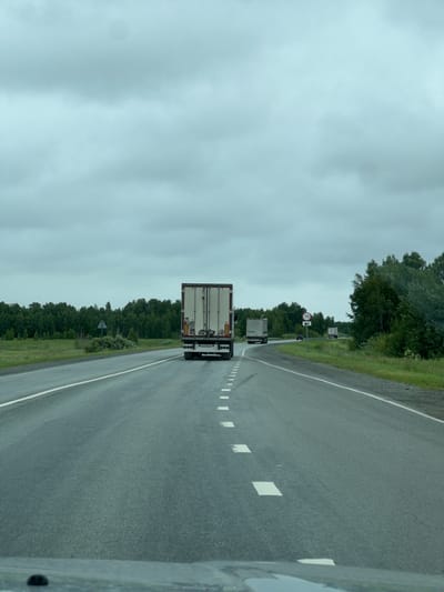 A vertical, medium-shot image captures a view from a vehicle traveling on a paved road. The road stretches ahead, bordered by a green, tree-lined shoulder on the left and a grey shoulder on the right. The sky is overcast and a pale blue-grey. The overall scene is somewhat dreary due to the cloudy weather.
In the foreground, the rear of a white semi-trailer truck is visible, its trailer a large, rectangular box. Further down the road, the trailers of two other semi-trailer trucks can be seen. One truck appears to be slightly ahead of the other. On the left side of the road, a circular traffic sign with the number "70" in red inside a white circle is mounted on a pole, indicating a speed limit. Another sign, possibly a warning sign for a bend or hazard, is visible amongst the trees.
The road has white lane markings that appear as dashed lines, with larger white squares marking intervals further down. The right edge of the road is partially obscured by what seems to be the side of the vehicle from which the photo is taken. The trees lining the road are dense and dark green. The time of day is indeterminate due to the overcast conditions, but it appears to be daytime. The location context is Kanashevo, Russia.