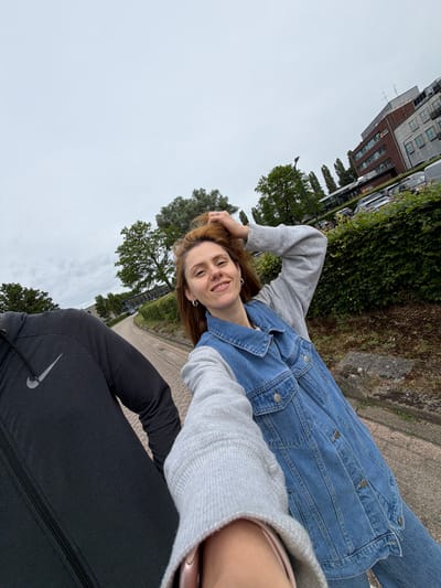This is a selfie taken outdoors in Son en Breugel, Netherlands. In the foreground, a woman with shoulder-length auburn hair and a denim jacket over a gray sweatshirt smiles at the camera, with her hand in her hair. To her left, a person wearing a black Nike hoodie is partially visible. The background shows a paved pathway, green hedges, trees, a parking lot with cars, and modern buildings. The sky is overcast.