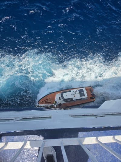 The image shows a lifeboat being lowered into the ocean from a larger vessel. The lifeboat is orange and white with solar panels on its roof. Two individuals, one wearing a yellow vest and the other in a blue shirt, are visible on the lifeboat, appearing to be involved in the operation. The ocean is a deep blue with white foamy waves. Part of the larger vessel, a white railing and blue deck, is visible in the foreground. The image is taken from a high vantage point, looking down onto the lifeboat and the water. The specific location in Santa Eulària des Riu, Spain, cannot be determined from the image alone.