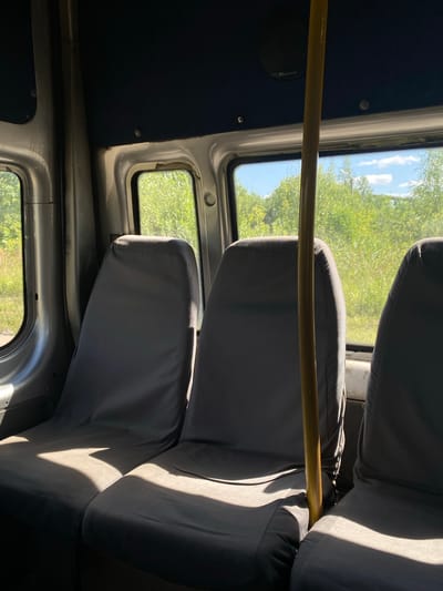 The image shows the interior of a passenger vehicle, likely a bus or van, with seats covered in dark grey fabric. Sunlight streams in through the windows, casting distinct shadows across the seats. A yellow pole, possibly a grab handle, stands vertically in the foreground between two seats. Outside the windows, lush green foliage and a blue sky with white clouds are visible, suggesting a rural or natural setting. The interior appears worn, with some of the metal framing and ceiling lining visible. The scene does not depict any people, specific city landmarks, or immediate actions. The location is stated to be Furmanov, Russia, though no visual cues within the image confirm this specific location.
