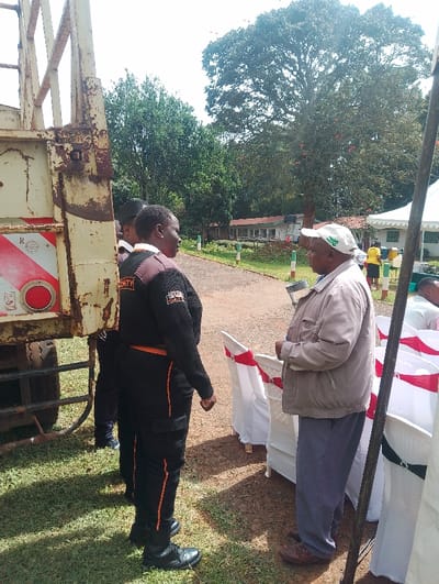 The image captures an outdoor scene, likely an event or gathering, in Gitaru, Kenya, under bright daylight.
In the foreground, two men are engaged in conversation. On the left, a man stands facing right, dressed in a dark security uniform featuring orange piping and the word "SECURITY" visible on the back of his vest. He wears black shoes. Facing him on the right is an older man in casual attire, consisting of a light-colored jacket, grey trousers, and a white baseball cap with an indistinct green logo. He wears brown shoes. Between them and to their right, a row of white chairs, neatly arranged and decorated with red sashes, suggests a formal seating area for an event. A white tent structure is partially visible on the far right.
To the left, a substantial, rusty metallic structure, possibly part of a truck or trailer, is prominent. It features red and white striped reflective markings and a circular red reflector. Green grass covers the ground where the men are standing.
In the mid-ground and background, a paved or dirt road curves gently into the distance. The area is lush with greenery, featuring numerous trees with full canopies, some displaying reddish flowers. In the far background, several single-story buildings with light-colored walls and dark roofs are visible, hinting at an institutional or community setting. Other individuals can be seen in the background, including one person in a yellow top near the buildings.
The bright, even lighting indicates it is daytime. The overall scene suggests a community event or function taking place in a natural, open environment.