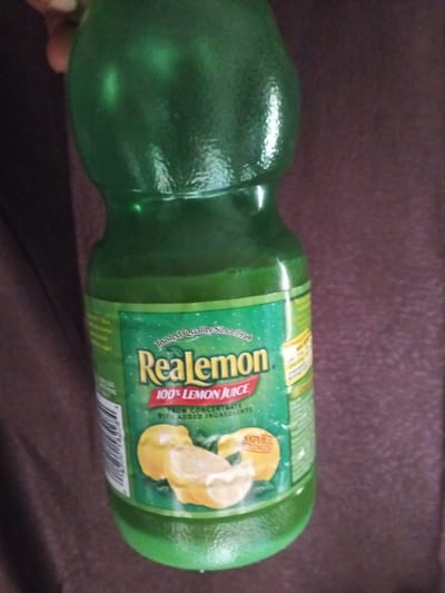 A green plastic bottle of Realemon 100% lemon juice is shown. The bottle has a textured surface and a green liquid inside. The label is predominantly green with yellow and red accents. The text "Realemon", "100% LEMON JUICE", and "Trusted Quality Since 1934" are visible. There are images of lemon slices on the label. The bottle is held by a person whose hand is partially visible on the left side of the image. The background is a dark brown, textured surface, possibly fabric. The scene appears to be indoors. There is no information in the image to suggest a location in Ashata, Nigeria.