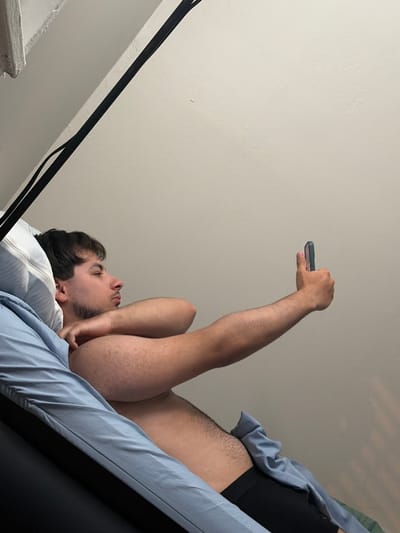 A young man is depicted lying down, possibly in a bed, looking upwards at a smartphone. He has dark hair and a beard, and his head rests on a white pillow, partially covered by light blue bedding. His right arm is extended, holding a dark-colored smartphone in a case, pointed towards the ceiling. He appears to be wearing dark underwear or shorts under the bedding on his lower body.
The man's expression is neutral and focused on the phone, indicating he is engaged with the device. The background consists of a plain, light-colored (off-white) ceiling or wall. A black strap or cable is visible running diagonally along the lower left edge of the frame, possibly part of a bed frame or furniture. The scene is indoors, in Franklin Township, United States of America. There are no specific time-of-day indicators or visible text in the image.