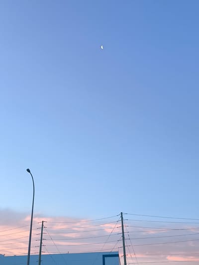 A crescent moon is visible in the clear, light blue sky above a city landscape. A tall, dark street lamp stands on the left side of the image. In the foreground, power lines are strung between utility poles, stretching across the frame. Below the power lines, a pale blue structure with a dark blue outline is partially visible. The lower portion of the sky features wispy clouds tinged with pink and orange, suggesting either dawn or dusk. The scene appears to be an outdoor urban environment in Mirabel, Canada, likely captured during twilight hours.