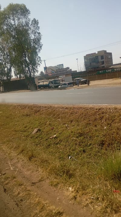 The image captures an outdoor urban or suburban street scene under clear, bright daylight conditions, likely late morning or early afternoon. The foreground features a strip of dry, light brown and green grass interspersed with patches of dirt, suggesting a somewhat arid environment or dry season. Some small pieces of litter, including a white object and a reddish item, are scattered on the grass.
A paved two-lane road runs horizontally across the midground, with a white line marking its edge. On the far side of the road, a developed area comes into view, bordered by a long green wall or fence. On this wall, the text "KY CAR WASH" is clearly visible in white lettering.
Behind the wall and further down the road, several vehicles are parked. These include a blue and white tanker truck, a light-colored car, and a darker SUV or pickup truck. Two individuals are visible near these parked vehicles, standing on the unpaved ground. One person is near the darker SUV, and another is slightly further to the left. Their activities are not discernible. To the far left, partially obscured by trees, another individual appears to be standing near a patch of disturbed ground, possibly engaged in an activity like landscaping, although this is unclear.
The background is dominated by several buildings. On the right, a modern multi-story building with a glass facade stands prominently. A sign on this building clearly reads "SALEM HOTEL." To the left of the hotel, another multi-story building with a colorful, patterned facade (featuring blue, green, and possibly orange/red sections) is visible. On this building, text reads "CACTUS APARTMENTS" with "RENTALS" below it. Further to the left, large leafy trees with slender trunks partially obscure additional structures. Utility poles and overhead wires crisscross the sky above the buildings. The sky itself is a uniform, clear pale blue, indicating good weather.