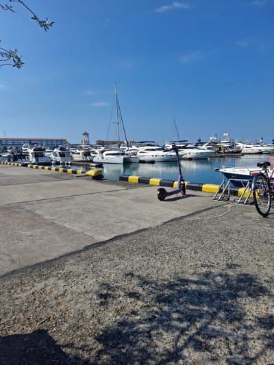 The image depicts a marina in Chvizhepse, Russia, on a clear, sunny day. In the foreground, a concrete pier with yellow and black striped bollards extends into the frame. A purple electric scooter is parked on the pier, to the right of which is a bicycle with a red seat. In the background, numerous white yachts are docked in the blue water. A building with a red roof and arched windows is visible behind the yachts. The sky is a clear, bright blue.