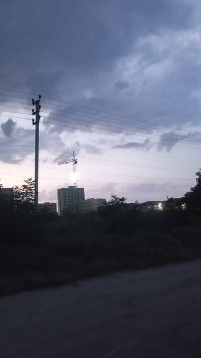 The image depicts an outdoor scene at dusk, likely in Baksan, Russia, characterized by a muted color palette and soft lighting. The sky is filled with dramatic, dark clouds, suggesting impending rain or the aftermath of a storm. A tall utility pole with wires stretches across the frame, anchoring the composition and hinting at the presence of infrastructure. In the middle ground, a construction site is visible, with several multi-story buildings under construction. A prominent crane stands over one of the buildings, its arm extended as if in operation, though the low light conditions make it difficult to discern any active work. The foreground is dominated by dark silhouettes of trees and foliage, obscuring the ground level and adding a sense of depth to the image. The overall mood is somber and quiet, with the fading light and cloudy sky contributing to a contemplative atmosphere. There is no visible text in the image.