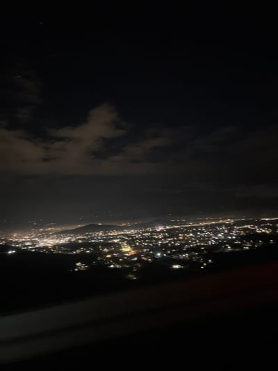 An aerial nightscape reveals a coastline speckled with bright city lights. The dense urban glow contrasts with the dark expanse of the ocean or sky. Some cloud cover is visible on the left. Part of the plane's interior frame can be seen on the right of the image.