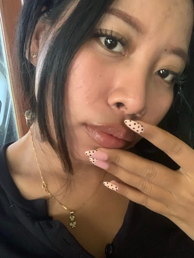 A close-up, chest-up, indoor portrait shows a young woman with dark hair and fair skin. She is wearing a black shirt and a gold necklace with a pendant shaped like two flowers. Her nails are decorated with a pink French manicure with black polka dots. She is touching her lips with her right hand. The background is out of focus, but appears to be a room with a wooden door frame. There is no visible indication of the city of Jajag, Indonesia.