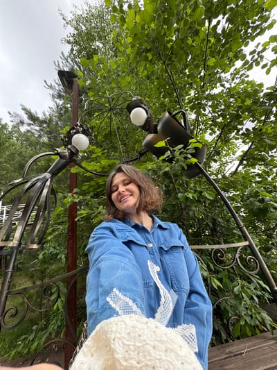 A medium shot captures a woman with brown hair, smiling broadly, as she takes a selfie outdoors. She is wearing a blue denim jacket and holding a white, crocheted item in her right hand, partially obscuring her lower face. Behind her, a metal sculpture resembling a human figure stands to her left, and a black, shovel-like object is visible to her right, both adorned with light bulbs. The background is dominated by lush green foliage and trees, with a cloudy sky overhead. The scene appears to be located in an outdoor seating area, possibly a park or garden, indicated by the presence of a wooden bench and ornate metal railings. There is no specific indication of the location being in the city of Kostovaty, Russia.