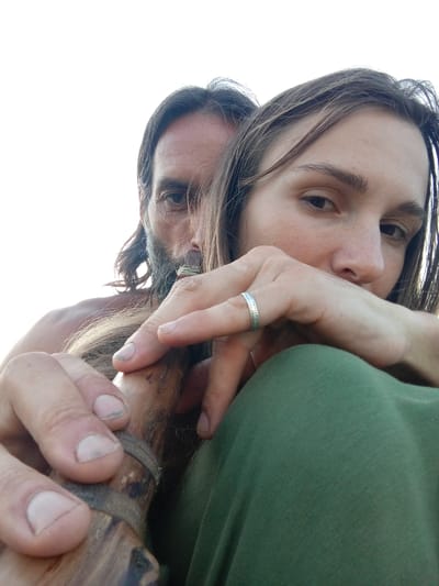 A low-angle, medium shot shows two individuals in an outdoor setting. The person in the foreground, facing the camera, is a woman with long, brown hair and fair skin. She is wearing a green garment. Her left hand, adorned with a silver ring, is resting near her face. Partially obscured behind her, a man with long, dark hair and a beard is visible. He appears to be holding a wooden object. The background is a bright, overcast sky.