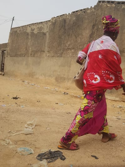A woman in vibrant, colorful West African clothing walks on a dirt path beside a weathered, grey concrete wall. The path is littered with trash and debris. The wall is worn and shows signs of age and disrepair. The location appears to be Dutsinma B, Nigeria. The woman wears a patterned headscarf, a red and white floral-patterned shawl, and a pink and yellow patterned skirt. She carries a beige bag over her shoulder. She wears flat, open-toed shoes. The sky is overcast and grey.