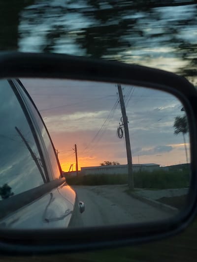 The image shows a car side mirror reflecting a sunset scene. In the reflection, there's a road stretching towards the horizon with utility poles and wires along the side. Vegetation borders the road. The sunset casts an orange glow. The mirror's frame is visible, with a blurred out-of-focus image of trees in the surrounding environment. The car's hood is also visible in the lower portion of the mirror. The scene is unlikely to be in Lukovit, Bulgaria, as the image appears to be taken in a rural area, and the city of Lukovit is a small, mostly urban city.