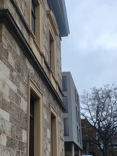 The scene depicts an upward-angled, low-light urban architectural view in Göttingen, Germany. The foreground features the upper two levels of a historic building located at Weender Tor 4, constructed with roughly cut, coursed stone blocks in variegated shades of grey and beige, exhibiting prominent mortar lines. Two vertically rectangular windows are visible, framed by detailed light-cream colored stucco surrounds; the upper frame incorporates an arched pediment motif. A dark grey horizontal cornice runs across the facade below the upper windows. The roof's dark grey soffit is partially visible at the top left.

To the right and set back, a modern, multi-story building identified as Am Weender Tor 2, with a smooth, light grey facade, presents a stark contrast. It features multiple narrow, vertically oriented dark windows or panels, arranged in a rectilinear pattern, and a flat roofline. In the background, a large, bare deciduous tree with a dense canopy of fine branches is visible, partially obscuring an older building with a reddish-brown tiled roof and a single round attic window. The sky is uniformly overcast and light grey, suggesting diffuse natural light. No individuals or discernible activities are present within the frame.