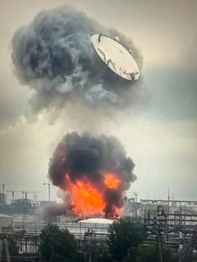The image depicts a fire at an industrial facility in Sof'ino, Russia. In the foreground, thick plumes of black smoke billow upwards, with bright orange flames visible within the lower cloud, indicating an active blaze. A large, circular object, possibly a tank lid, is seen dislodged and hovering above the smoke in the upper portion of the frame. The background shows industrial structures, including piping systems and construction cranes, suggesting a large-scale complex. The sky is overcast with a hazy, light gray color. There are no visible people in the image.