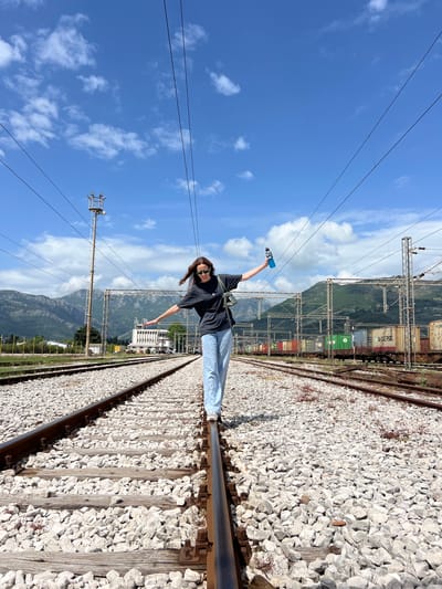 A young woman stands on a train track near Polje, Montenegro. She balances on the rail, arms outstretched. She holds a blue bottle in one hand and wears sunglasses, a black t-shirt, and blue jeans. The train track runs diagonally across the frame, with white gravel on either side. In the background, mountains rise behind shipping containers on a parallel track. Utility poles and wires stretch across a blue sky with scattered clouds. Buildings are seen in the distance.