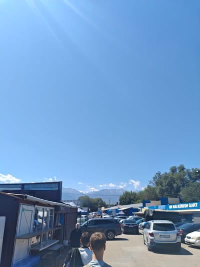 The image depicts an outdoor market scene in Іргелі ауылдық округі, Kazakhstan. The sky is a clear, bright blue with sun rays visible. In the foreground, there are several people, two of whom are walking away from the camera. To the left, there's a small market stall or kiosk with a brown metal exterior and a white framed window. Behind this stall, more market structures and vehicles are visible. In the mid-ground, a row of parked cars lines a paved area. Further back, trees and a large blue and white building with signage that reads "UN MAI KURISH QANT" are present. In the distance, mountains can be seen under the blue sky. The overall impression is a busy outdoor market on a sunny day.