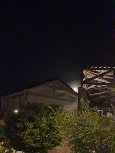 The image captures a nighttime scene in Charnay-lès-Mâcon, France. A brightly lit moon is visible through a gap between two buildings. The building on the left appears to be made of stone and timber, with a pitched roof. In front of this building, there is lush green foliage. To the right, a large wooden structure with a diagonal bracing pattern is prominent. The sky is dark, indicating it is night. No people are visible in the frame.