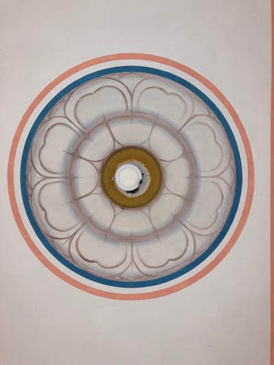 The image shows a circular ceiling fresco with a lotus flower motif. The fresco is painted in shades of grey and brown, with outer rings of blue and coral pink. In the center of the fresco, a modern white LED light fixture is installed within a yellow circular border. The background is a plain white or off-white surface, likely a ceiling. The image was captured in Sri Jayewardenepura Kotte, Sri Lanka, likely in a temple or a similar religious or decorative building.