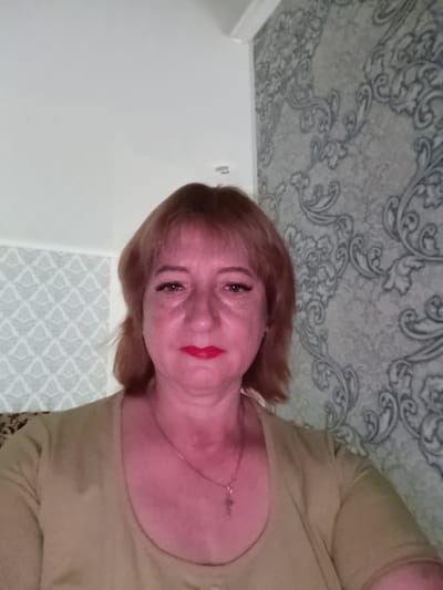 A medium-shot, front-facing selfie shows a middle-aged Caucasian woman with shoulder-length auburn hair. She is wearing a tan, long-sleeved shirt and a silver necklace with a small cross pendant. The woman has fair skin, noticeable freckles, and is wearing red lipstick and eyeliner. Her expression is neutral. The background consists of a wall with ornate grey and white wallpaper on the right side and a lighter patterned wallpaper on the left. A section of white ceiling is visible at the top. The lighting in the image is reddish, suggesting it might be artificial light or a color filter. The image appears to have been taken indoors, possibly in a residential setting in Tugulym, Russia.