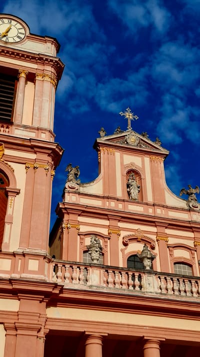 The image shows a close-up, low-angle view of the ornate facade of the Göttweig Abbey in Furth bei Göttweig, Austria, against a vibrant, cloudy blue sky. The architecture is Baroque, characterized by its pink and cream color palette, gold embellishments, and detailed sculptural elements.
On the left side of the frame, a portion of a tower is visible, featuring a large clock face with Roman numerals and a gold hand pointing to approximately 11:55. Fluted columns and decorative moldings are prominent.
The central and right portions of the image focus on the main facade, which is adorned with statues. Two winged angelic figures flank a pedimented section containing a niche with a statue of a Madonna and child, holding a scepter. Above this niche, a gilded sunburst cross crowns the building.
A stone balustrade runs across the mid-section of the facade, supported by pillars and decorated with finials. Smaller sculpted figures and urns are positioned on pedestals above and around the windows. The lower part of the facade features large, rounded columns and intricate cornices. The lighting suggests it is daytime, with strong shadows cast by the architectural elements. There are no people visible in the image.