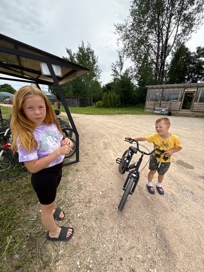 In Zamosh'e (Zaklinskoe S/P), Russia, two children are present outdoors on a cloudy day. To the left, a young girl with long, reddish-blonde hair stands facing the camera, wearing a purple tie-dye t-shirt with graphics and black shorts. She has on black slide sandals. Behind her, a rack of bicycles is visible under a dark-framed shelter with a translucent roof. To the right, a young boy in a yellow t-shirt with graphics and camouflage shorts is holding a black bicycle. He is wearing white socks and dark clogs with red and white star patterns. The ground is a mix of dirt and gravel. In the background, there is a wooden building with glass windows and trees.