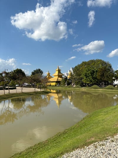A golden temple is reflected in the murky water of a pond. Lush green grass and a path line the water's edge. Tall trees partially obscure the temple. A blue sky with puffy white clouds fills the upper left corner.