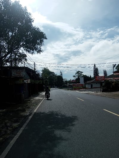 The scene depicts a daytime view of a paved two-lane road in Domphoog_Dungkarchhoeling, Bhutan, under a sky with scattered white clouds. The road, marked with a dashed yellow center line and a solid white line on its left edge, recedes into the distance. On the left side, a large, dark-foliaged tree casts a prominent shadow across the road. Beneath the tree, a building is partially visible, featuring a sign with the word "REPLEKSI" legible on it. Numerous utility wires are strung horizontally across the upper-left and central portions of the frame. A single person, wearing a dark helmet and clothing, operates a motorcycle, traveling away from the viewer in the left lane. On the right side of the road, several buildings are visible, one prominently displaying a red sign with white text that reads "TAHU SUMEDANG". Coniferous and deciduous trees are interspersed among the buildings and along the roadside.