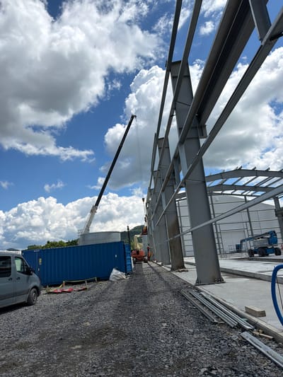 A construction site is depicted in Stróża, Poland, under a partially cloudy blue sky. In the foreground, a grey van is parked on a gravel surface, to the left of a large blue shipping container. Several pieces of metal scaffolding and a white bag are also on the ground near the container. The main focus of the image is the skeletal structure of a large building under construction, composed of grey steel beams. A tall mobile crane with a black boom is positioned behind the shipping container, with its arm extended upwards. Further in the background, an orange construction vehicle is visible, as well as a blue aerial work platform. A lighter-colored, cylindrical structure with a flat top stands behind the blue container. The environment suggests an active construction project with materials and machinery present.