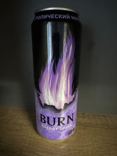 A tall, purple and black energy drink can is displayed upright on a wooden surface. The can features a stylized flame design in shades of purple and white. Text on the can reads "BURN ENERGY DRINK" and "449 ml." The top of the can has text in Russian, which appears to be "ТРОПИЧЕСКИЙ МИКС" (Tropical Mix). The background is a dark, out-of-focus wall. The scene is lit from the front, casting a shadow behind the can. No people are present, and the location is not identifiable as being in Tambov, Russia, or any specific city.