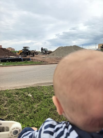 The image depicts a construction site in Froli, Russia. In the foreground, a blurry close-up of a person's head is visible, wearing a striped shirt. To the left, a white baby stroller is partially seen. The middle ground shows a road running horizontally across the frame. Beyond the road, construction activities are underway. Two yellow and black bulldozers are present; one is digging into a pile of gravel, and another is parked. A large mound of gravel dominates the right side of the frame. Piles of dirt and black pipes are also visible. Buildings and utility poles are scattered in the background under a cloudy sky.