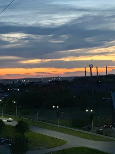 This is a landscape photograph taken from an elevated vantage point, likely a building. The image is oriented vertically, showing a scene at dusk or dawn.

The left side of the frame is dominated by a dramatic sky filled with wispy clouds. These clouds are illuminated by the setting or rising sun, creating a gradient from deep blue-grey on the far left to vibrant oranges and yellows closer to the horizon.

The right side of the image depicts a semi-urban or industrial landscape. In the middle ground, several tall industrial chimneys or cooling towers are visible, silhouetted against the colorful sky. Beyond these, there is a line of buildings with visible windows, suggesting a factory or residential complex. Further into the distance, a scattering of lights indicates a town or city.

The foreground on the right shows a paved road with a grassy verge. Several streetlights are visible along the road, some illuminated, contributing to the dim lighting of the scene. A few cars are parked or driving along the road. Dark trees and shadows suggest the presence of foliage.

The time of day is clearly indicated by the sky's colors and the presence of artificial lighting in the developed areas. The weather appears to be clear, allowing for a striking sunset or sunrise. The overall mood is one of transition, with the natural beauty of the sky contrasting with the man-made structures and infrastructure. There is no visible text in the image.
