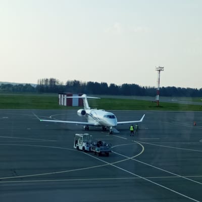 An aerial view captured from a high vantage point shows a modern, silver private jet parked on an airport tarmac. The jet has its distinctive swept-back wings and is positioned centrally in the frame, facing slightly to the left. In front of the jet, a small white tug vehicle is attached to a baggage trailer, which appears to be loaded with luggage. Two individuals in high-visibility vests are standing near the jet, possibly ground crew attending to the aircraft. The tarmac is marked with white lines, indicating taxiways and parking areas. In the background, a red and white striped building is visible, along with a tall, striped airport beacon or mast. Beyond the tarmac, a green grassy field stretches towards a line of trees under a hazy, pale sky. The image appears to be taken in the city of Naberezhnye Chelny, Russia, given the context.
