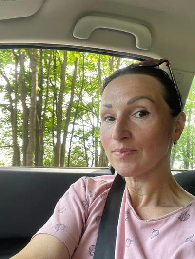 A woman with dark hair is taking a selfie in the back seat of a car. She is wearing a pink t-shirt with a unicorn pattern and a seatbelt is visible. Sunglasses are perched on top of her head. The background shows trees and foliage seen through the car window, suggesting a natural setting outside the city of Zelenogorsk, Russia. The interior of the car, including the headrest and a handle, is also visible.