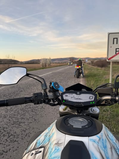 The image depicts a motorcycle's point-of-view shot on a paved road near Belanitsa, Bulgaria. The motorcycle's dashboard indicates a speed of 0 km/h and a time of 8:09. The fuel tank has a white and grey design with blue lightning bolts and a playing card of the Ace of Spades. Another motorcycle is visible in the distance on the road ahead. A road sign is visible on the left side of the road, adjacent to a grassy area. The sky is light with hues of yellow, suggesting it is either sunrise or sunset.