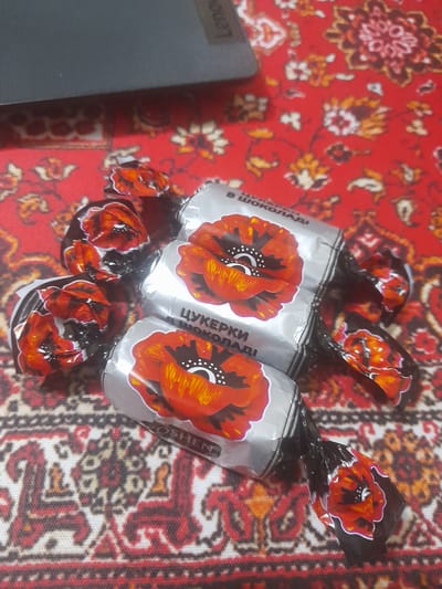 The image shows three Roshen chocolates wrapped in silver foil with a red poppy flower design, laying on a red patterned textile that might be a rug or tablecloth. The chocolates are arranged close together, suggesting they might be grouped. A part of gray laptop is located in the left side of the image. The location is not discernible.