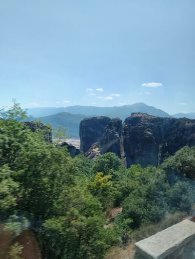 The image captures a panoramic view of the Meteora rock formations near Kalambaka, Greece. The foreground is dominated by lush green trees, some appearing slightly blurred due to motion, suggesting the photo was taken from a moving vehicle. In the mid-ground, imposing, dark-colored rock pillars rise dramatically from the landscape. These geological formations are characterized by their sheer, vertical cliffs and rugged texture.
Beyond the prominent rock pillars, a valley is visible with a scattering of buildings suggesting a settlement or part of the town of Kalambaka. The background is filled with layers of mountains fading into a hazy blue, indicative of distance. The sky above is a clear, bright blue with a few scattered white clouds. The overall impression is one of natural grandeur and geological wonder, characteristic of the Meteora landscape.