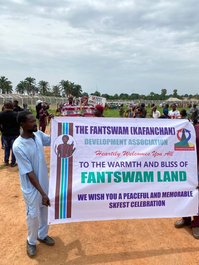 A man in a light blue shirt and dark pants holds up a banner that reads "The Fantswam (Kafanchan) Development Association Heartily Welcomes You All To The Warmth and Bliss of Fantswam Land. We Wish You A Peaceful and Memorable SKFEST Celebration." The banner is being held up in a field surrounded by trees and people. This is in Kafanchan, Nigeria.
