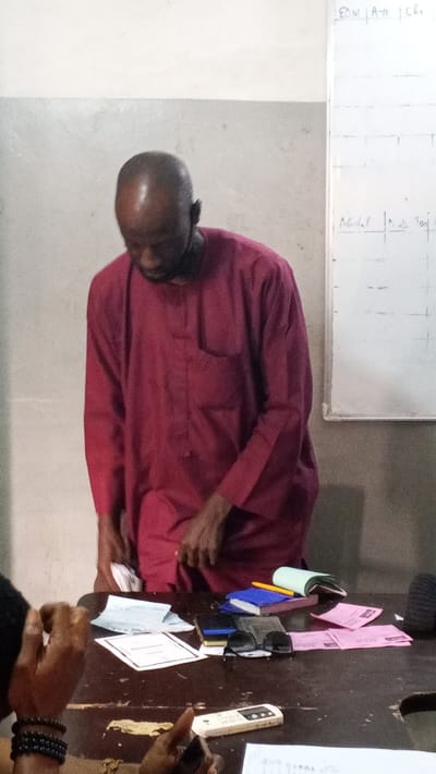 The scene is an indoor setting in Dakata, Nigeria, possibly a classroom or meeting room. A dark-skinned, bald adult male, dressed in a maroon long-sleeved garment, stands partially obscured, leaning forward and looking down towards a wooden table. His hands are positioned above items on the table. To the left, portions of at least two other individuals are visible, seated at the table. One person's arm, adorned with black beaded bracelets, rests on the table. Another person's hand holds a white remote control and a white smartphone. The wooden table surface is cluttered with various items, including white papers/documents (one appearing to be a certificate), several small pink paper slips, a small blue book, a yellow pencil, black sunglasses, and a dark wallet or small bag. In the background, a light-colored wall features a whiteboard with partially legible handwritten text, including "EDUI A-70", "CHR", "Nodal", and "N.b Twi". The overall arrangement suggests a gathering or administrative activity.
