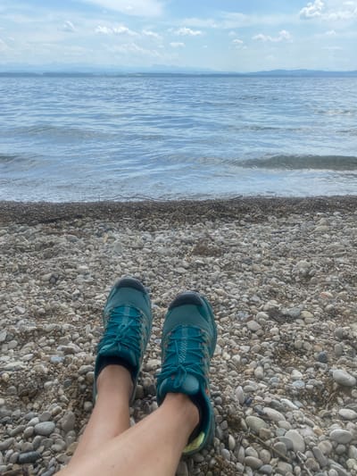 The image shows a view of a pair of teal running shoes on a pebble beach next to a body of water. The water extends horizontally to the left, with a line of dark pebbles along the edge closest to the shoes. The water appears calm with some ripples on the surface. Beyond the water, there is a faint horizon line under a light blue sky with some scattered clouds. The shoes are positioned in the lower right portion of the frame. The legs of the person wearing the shoes are visible, suggesting the perspective is from the person standing on the beach. Given the context of Cortaillod, Switzerland, the body of water is likely Lake Neuchâtel.