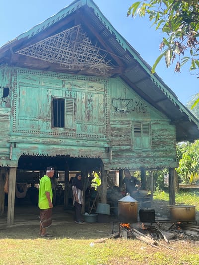 This image depicts an outdoor scene in the village of Pulo, Indonesia, centered around a traditional stilt house with weathered, teal-painted wooden walls. The house features intricate latticework under the gabled roof and decorative carvings on its facade. In the foreground, several large pots are suspended over open fires, suggesting food preparation. A man in a bright green shirt and a sarong stands to the left, facing away from the viewer, near the entrance of the house. To his right, a woman wearing a hijab and a striped top is seen looking towards the cooking area. Another man, partially obscured, is tending to the fires. The ground is covered in grass and dry leaves, and leafy branches of a tree hang over the top right of the frame, casting dappled shadows. The overall atmosphere is that of a rural village setting, possibly during a communal gathering or festival.