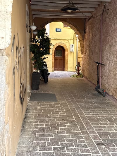 The image depicts a narrow, cobblestone alleyway in Chania, Greece. The alley is flanked by stone walls, one of which is painted a pale yellow and covered in graffiti. The other wall is rough-hewn stone. An arched entryway is visible in the distance, leading to a yellow building with a wooden door. A street sign with Greek lettering, reading "ΟΔΟΣ ΒΕΝΕΡΗ" (Odos Venere - Venere Street), is mounted above the entryway.
In the foreground, to the left, is a black motorcycle parked next to a dark planter with a leafy green tree. To the right, a red fire hydrant with coiled green hoses is situated. Further down the alley, an electric scooter stands upright against the stone wall. The cobblestone pavement is laid in a diagonal pattern with a contrasting strip running down the center. Overhead, exposed wooden beams support a section of the alley. A street lamp with two globes is attached to the yellow wall on the left.