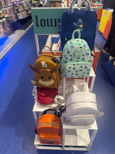 The image shows a retail environment, likely a store, featuring a display of character-themed backpacks. The backpacks are arranged on white shelving units. In the foreground, on the lower shelf, is an orange backpack with an embossed character design, possibly from the Pokémon franchise. Above it, on the middle shelf, sits a tan backpack with Eevee's face, ears, and tail prominently displayed, along with a small white flower charm and a Loungefly tag. To the right of the Eevee backpack is a white backpack with zipped compartments and what appear to be small, white wings. Further back on the middle shelf, a light blue backpack is adorned with Stitch and other characters from Disney's Lilo & Stitch. To the left of the blue backpack is another, larger blue backpack with a Stitch applique. In the background, shelves are stocked with various items, and a sign with "loun" in stylized lettering is visible. The floor is a speckled blue. The overall impression is a merchandise display, likely in a specialized store catering to fans of anime and pop culture. The question about Leers, France is not directly answerable from the visual information present in the image.
